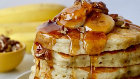 Stack of Banana Nut Pancakes topped with fresh banana slices, pecans and maple syrup; bananas and small bowl with pecans in background