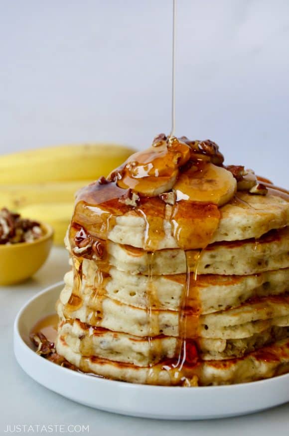 Stack of Banana Nut Pancakes topped with fresh banana slices, pecans and maple syrup; bananas and small bowl with pecans in background