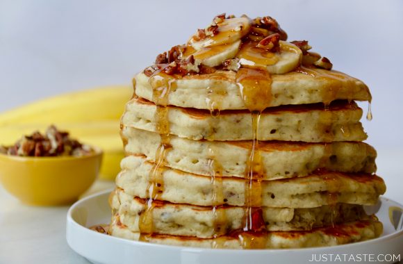 Tall stack of Banana Nut Pancakes topped with sliced fresh bananas, pecans and maple syrup on white plate with small bowl filled with pecans and bananas in background