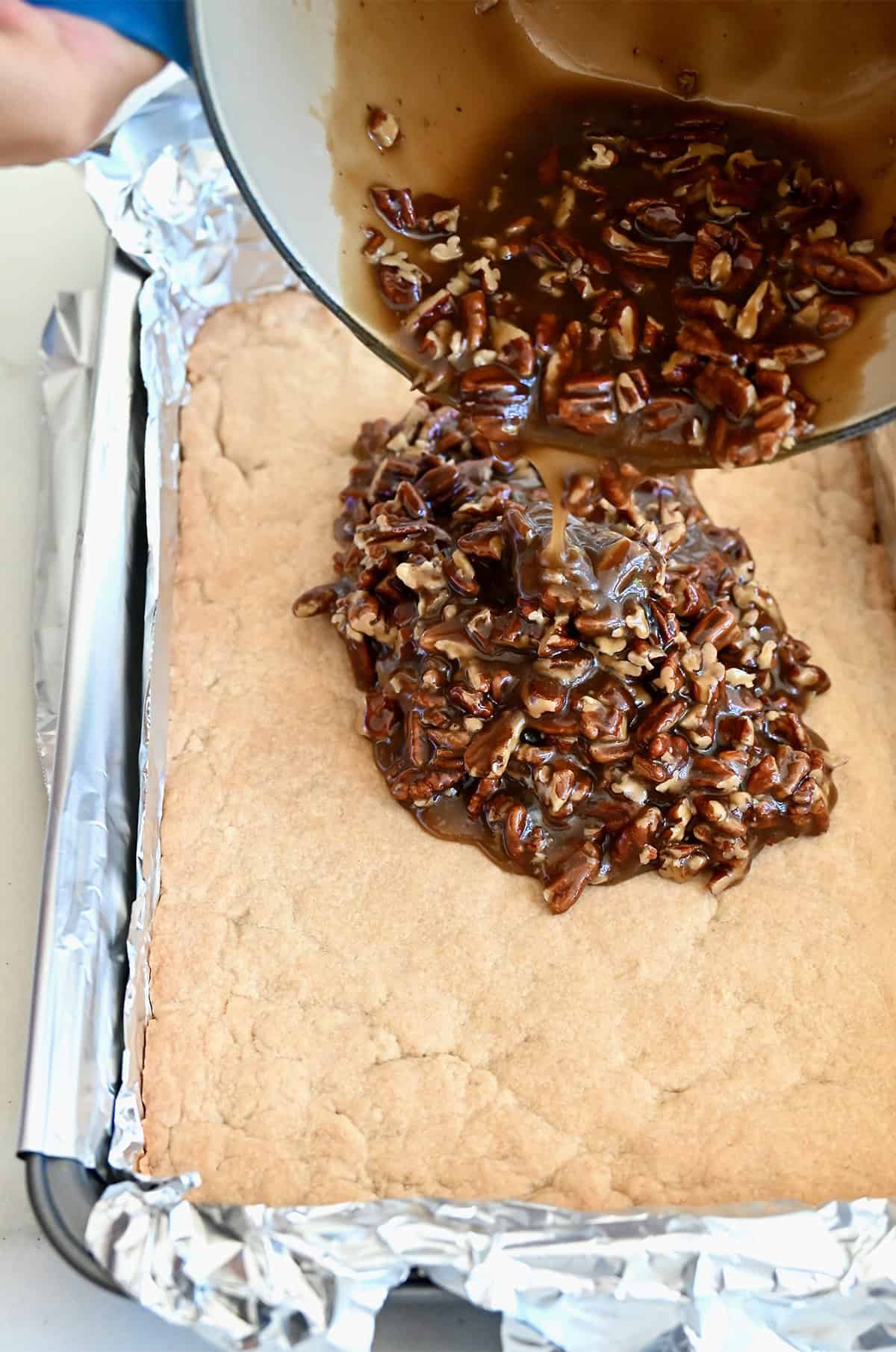 Pouring warm pecan pie filling atop parbaked shortbread crust in a tinfoil-lined 9x13 baking pan.