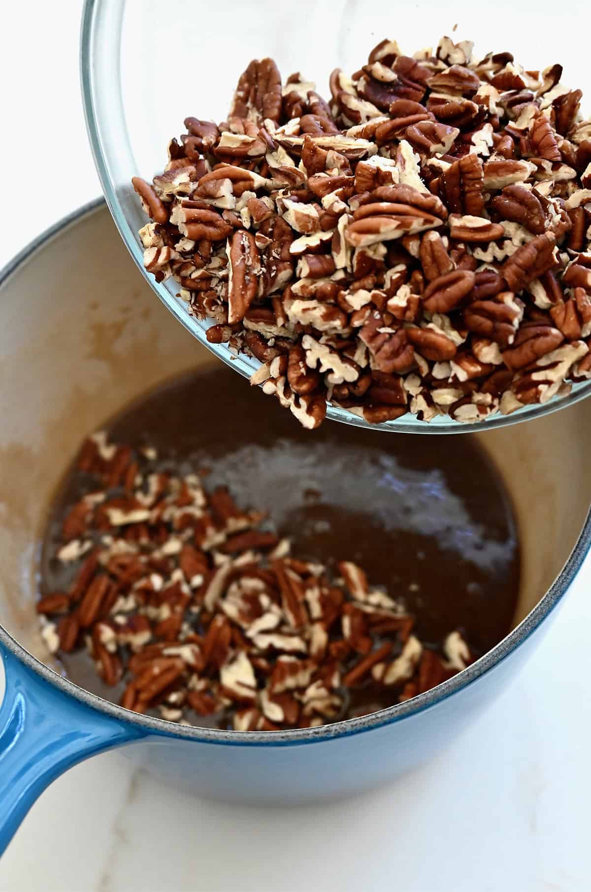 Chopped pecans are being added to a saucepan containing honey, brown sugar and heavy cream to make pecan pie filling.