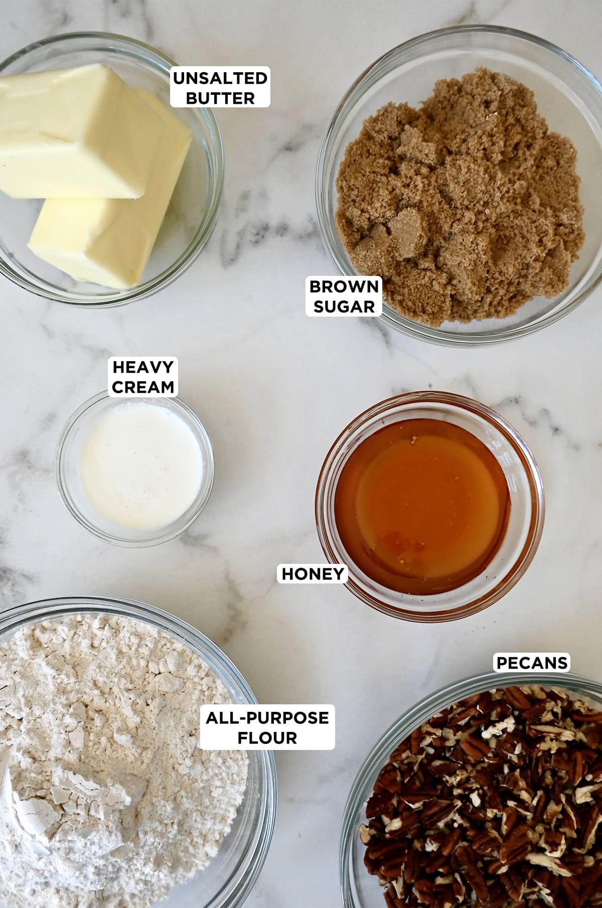 Ingredients to make pecan pie bars in glass bowls, including butter, brown sugar, heavy cream, honey, chopped pecans and flour.