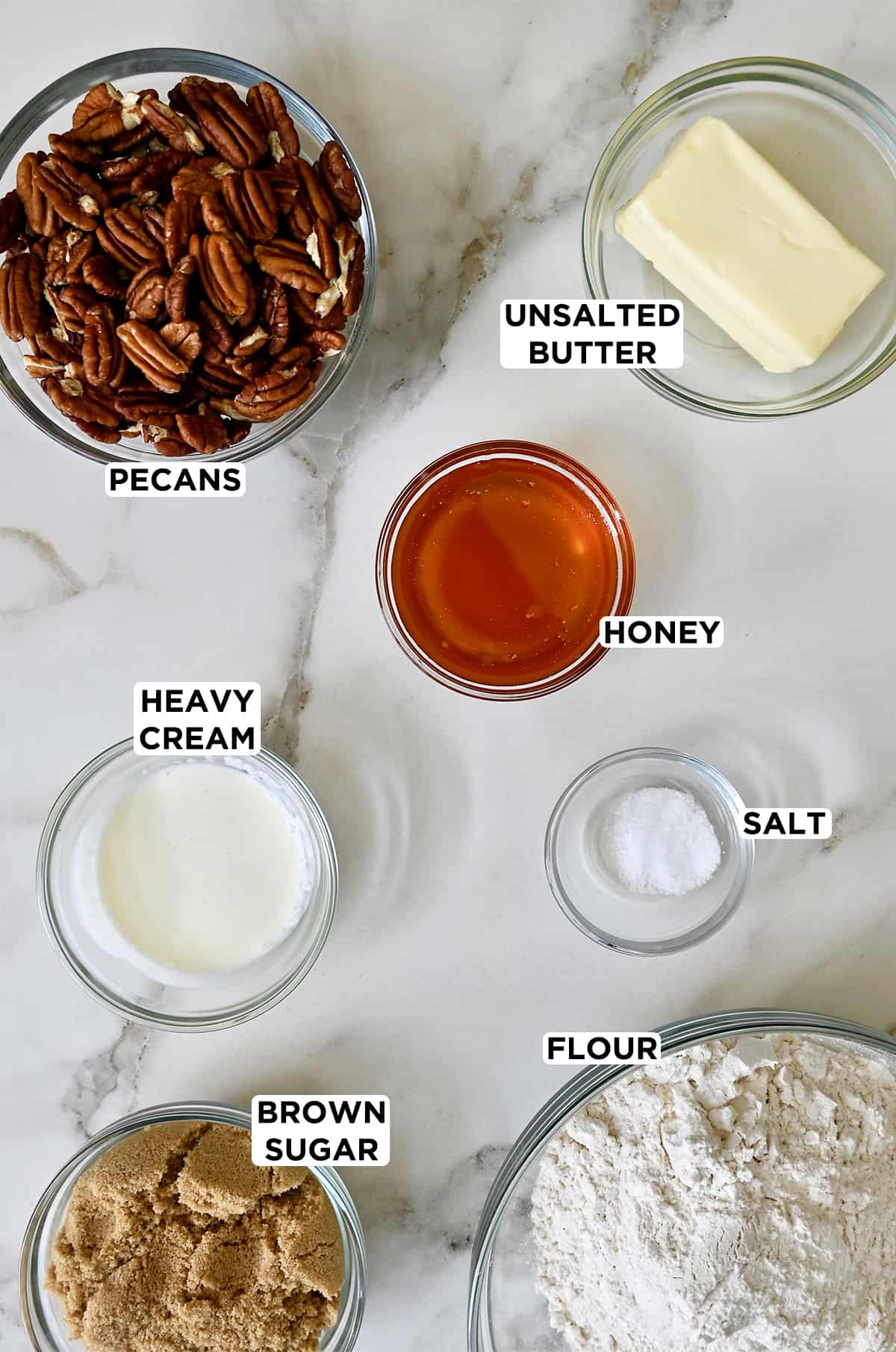 Various sizes of clear bowls containing halved pecans, sticks of butter, honey, salt, flour, brown sugar and heavy cream.