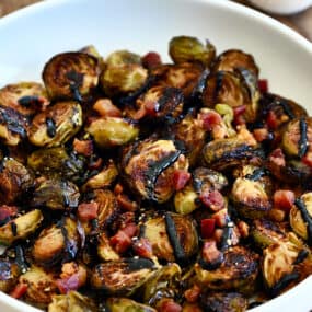 Roasted Brussels sprouts with pancetta bits all drizzled with thick balsamic syrup in a white serving bowl.