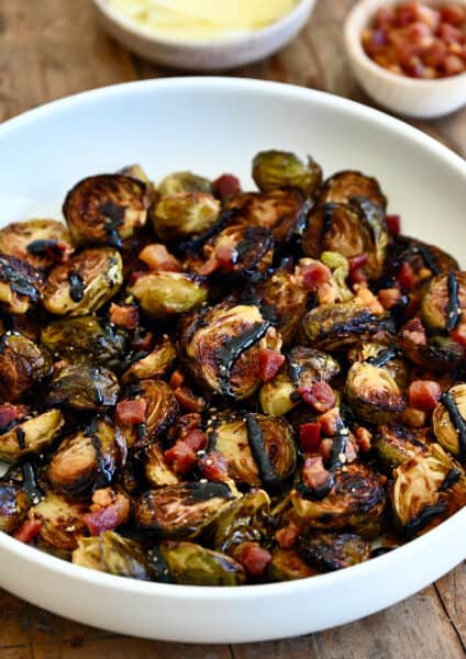 Roasted Brussels sprouts with pancetta bits all drizzled with thick balsamic syrup in a white serving bowl.