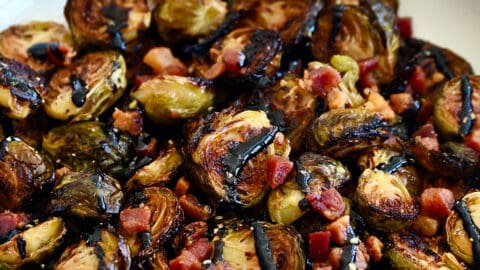 Roasted Brussels sprouts with pancetta bits all drizzled with thick balsamic syrup in a white serving bowl.