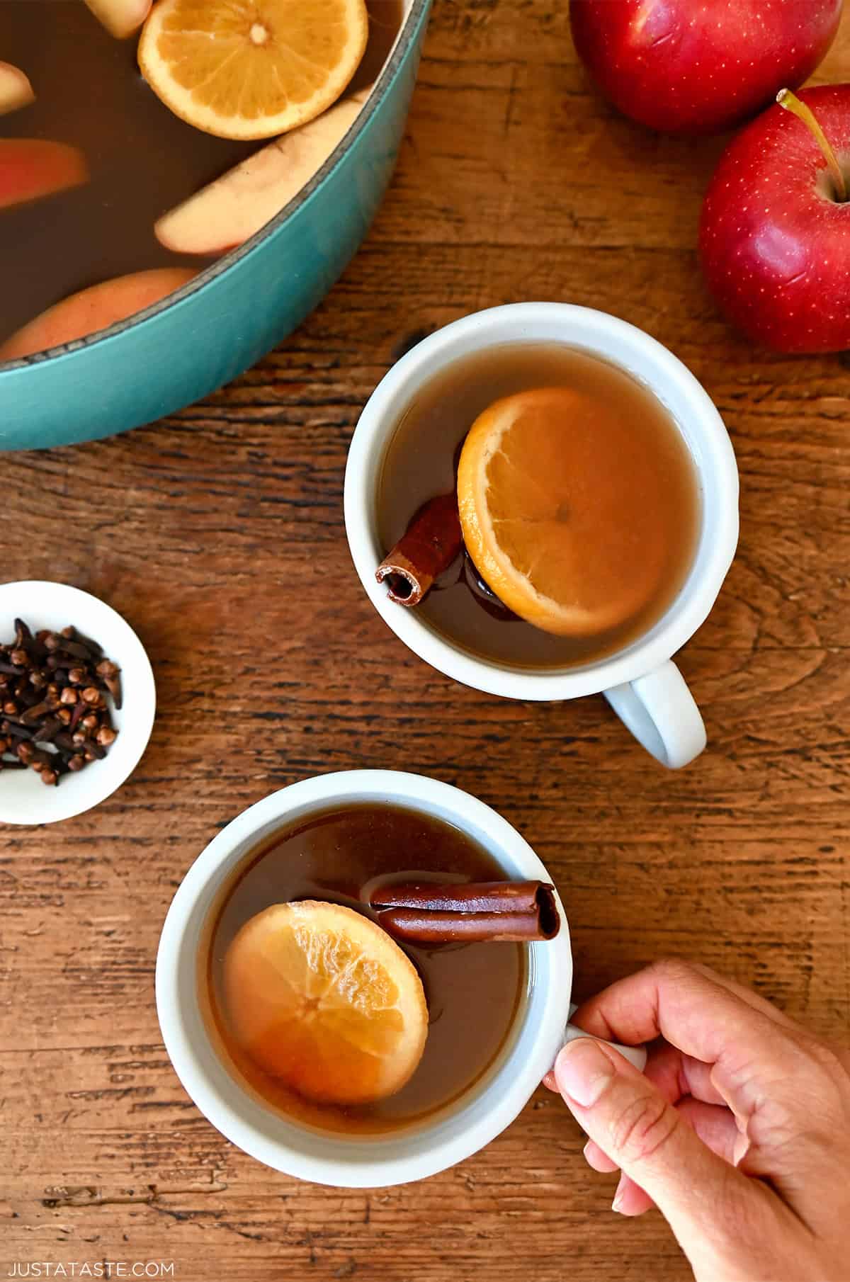 Two mugs filled with spiked apple cider garnished with a cinnamon stick and an orange slice. A small bowl with whole cloves and a pot with more mulled cider is nearby.