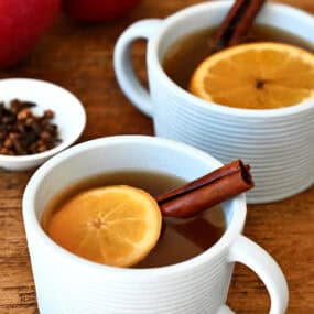 Two mugs containing spiked apple cider with an orange slice and a cinnamon stick.