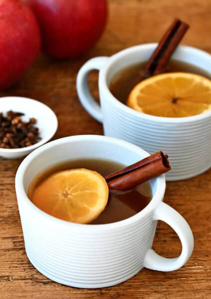 Two mugs containing spiked apple cider with an orange slice and a cinnamon stick.