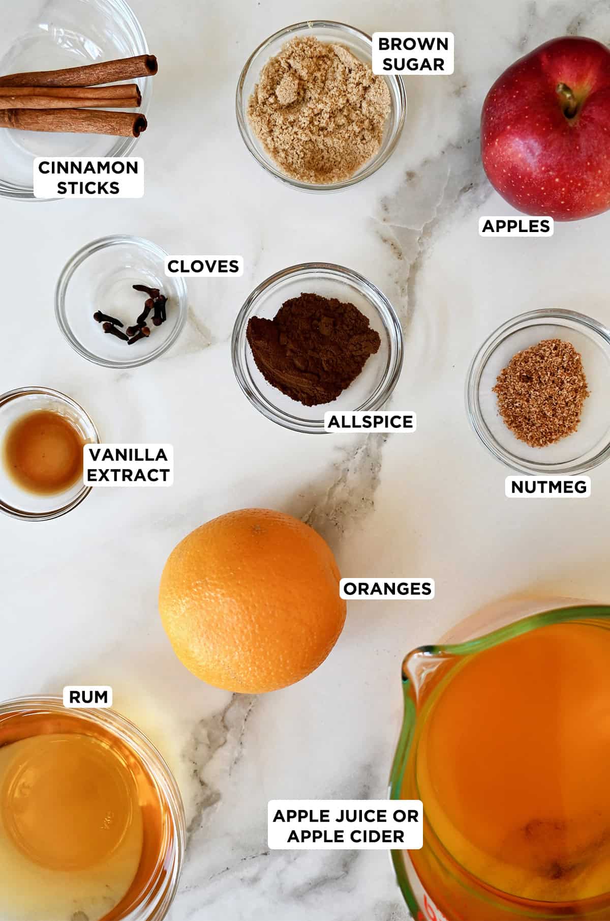 An orange and an apple next to various sizes of glass bowls containing cinnamon sticks, nutmeg, allspice, cloves, vanilla extract, rum, apple cider and brown sugar.
