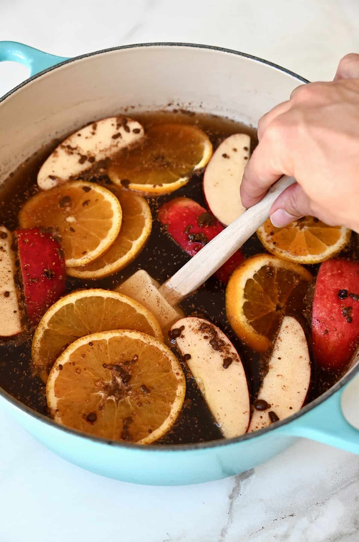 A hand holding a spatula stirs warm mulled cider in a large pot.