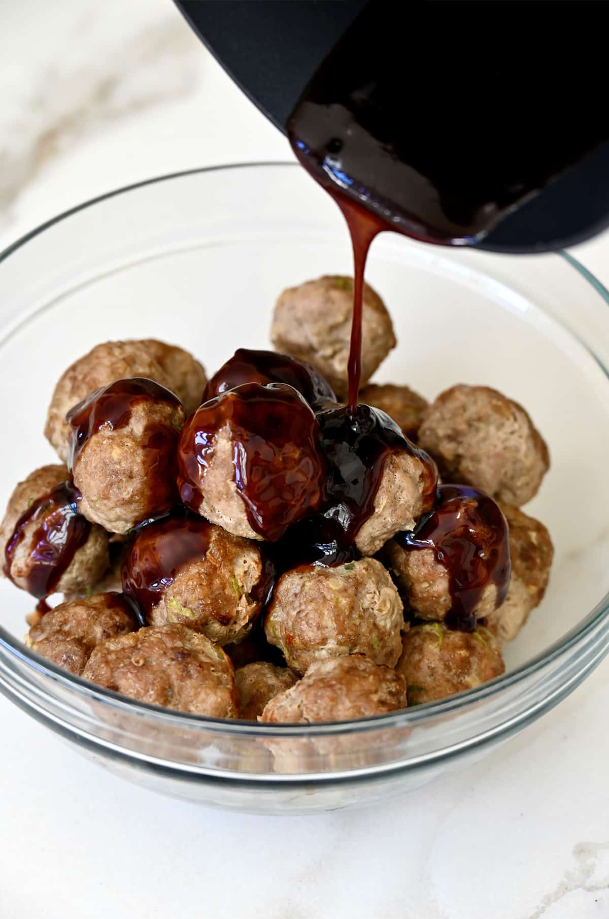 Blackberry glaze being poured over cooked meatballs in a clear glass bowl.