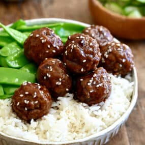 Blackberry jam meatballs garnished with sesame seeds atop white rice in a bowl with snap peas.