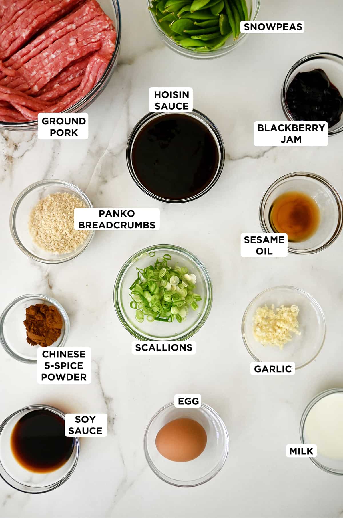 Various sizes of clear bowls containing the ingredients needed to make blackberry meatballs, including hoisin sauce, ground pork, sliced scallions, blackberry jam, sesame oil, garlic, egg, panic breadcrumbs, Chinese 5-spice powder, soy sauce and hoisin sauce.