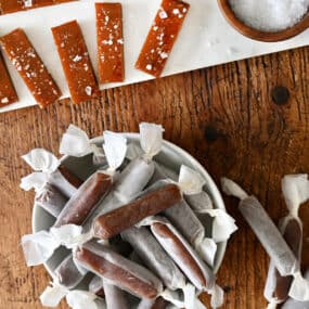 Homemade salted caramels wrapped in wax paper in a bowl next to more caramels topped with large-flake sea salt.