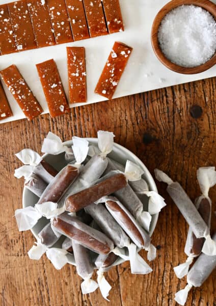 Homemade salted caramels wrapped in wax paper in a bowl next to more caramels topped with large-flake sea salt.