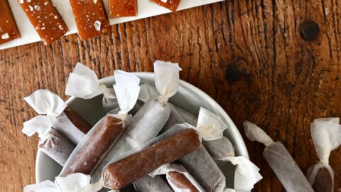 Homemade salted caramels wrapped in wax paper in a bowl next to more caramels topped with large-flake sea salt.