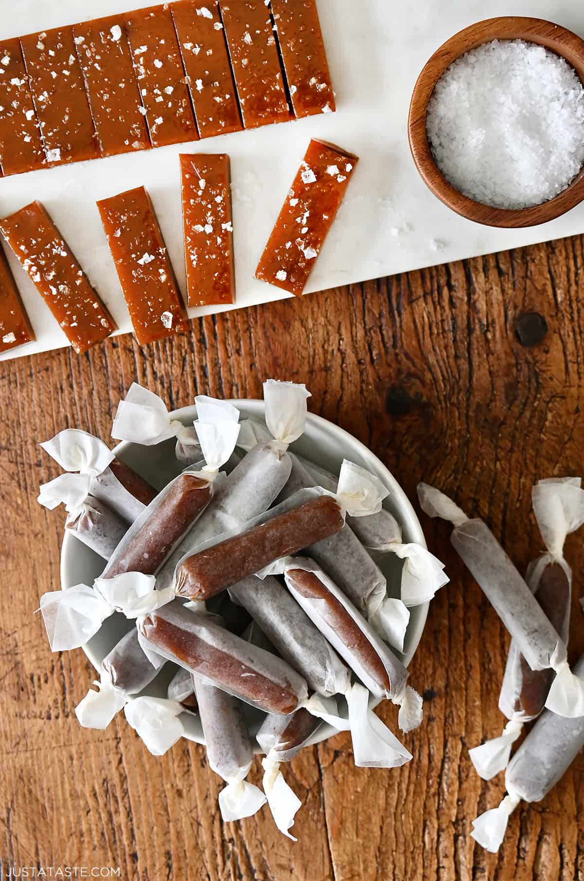 Homemade salted caramels wrapped in wax paper in a bowl next to more caramels topped with large-flake sea salt.