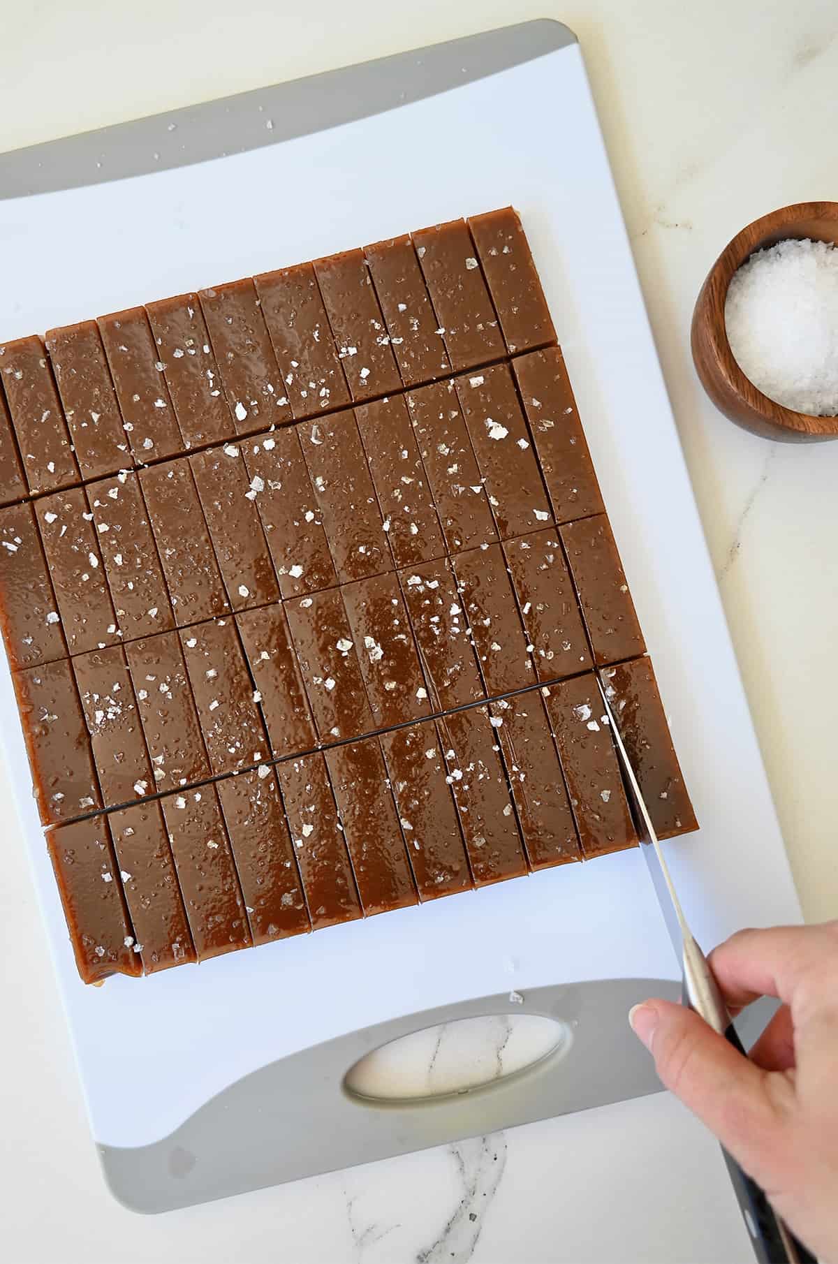 A hand holding a knife cuts into a block of soft caramels on a cutting board.