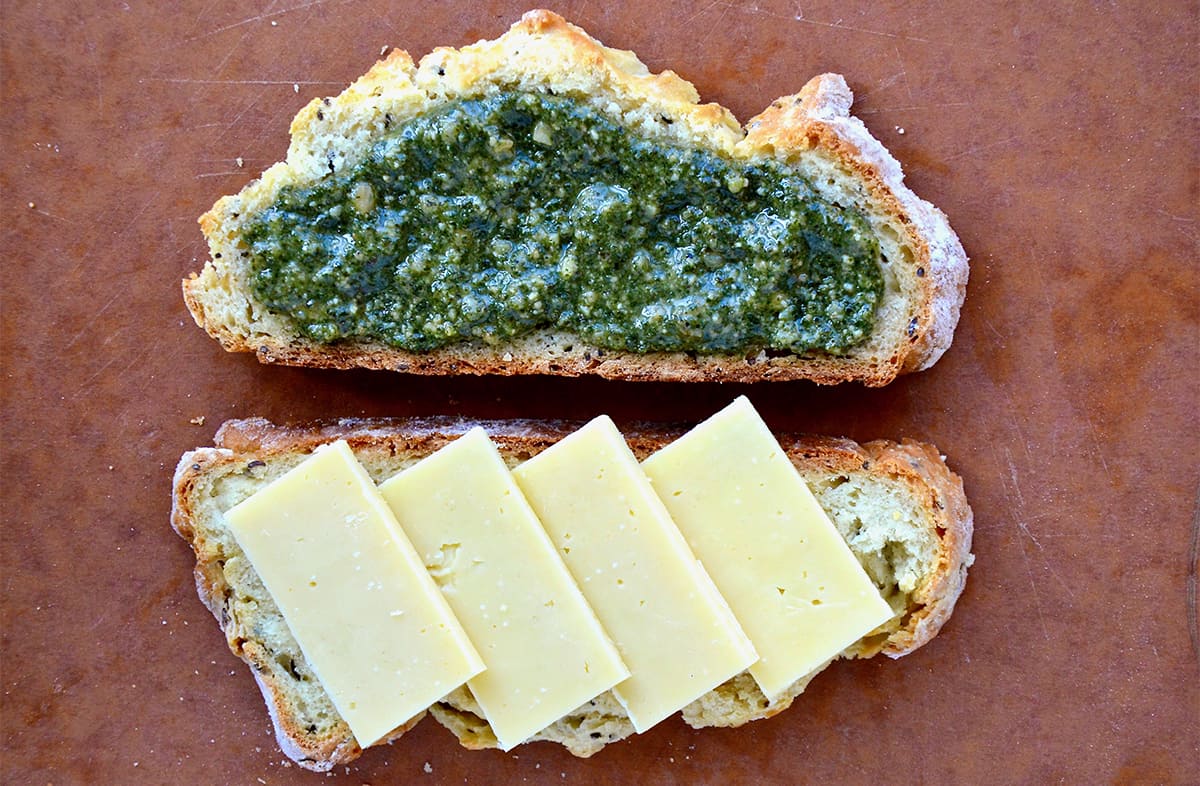 A slice of soda bread topped with basil pesto next to another slice of soda bread topped with four slices of cheese.