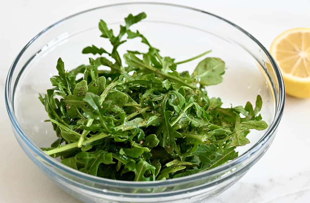 Fresh arugula with lemon juice, salt and pepper in a glass bowl with half a lemon nearby.