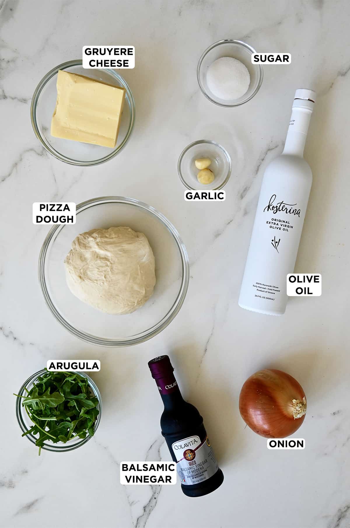 A bottle of Kosterina extra virgin olive oil and a bottle of balsamic vinegar next to a yellow onion and glass bowls containing a block of gruyere cheese, sugar, minced garlic, pizza dough and arugula.