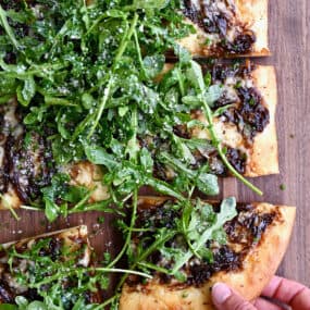 A hand reaches for a slice of French onion pizza topped with olive oil-dressed arugula.