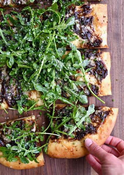 A hand reaches for a slice of French onion pizza topped with olive oil-dressed arugula.