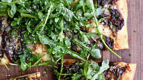 A hand reaches for a slice of French onion pizza topped with olive oil-dressed arugula.