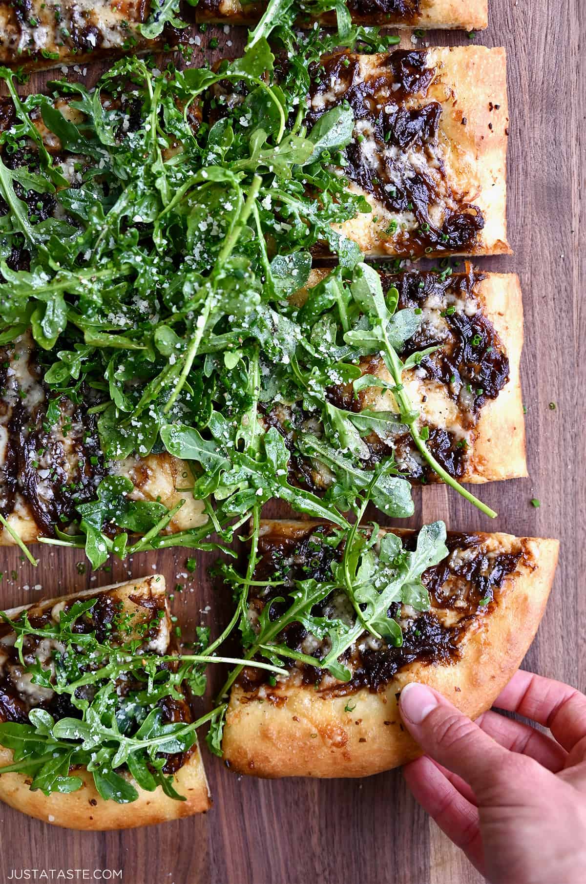 A hand reaches for a slice of French onion pizza topped with olive oil-dressed arugula.