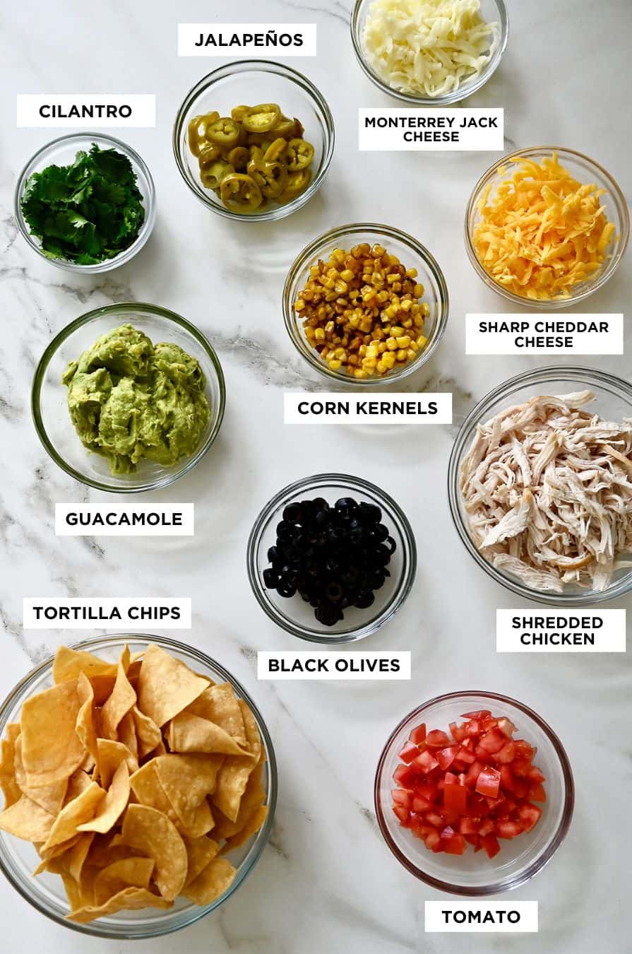 A top-down view of various sized bowls containing ingredients and labels for nachos, including tortilla chips, guacamole, cilantro, jalapeños, shredded cheese, corn kernels and shredded chicken.