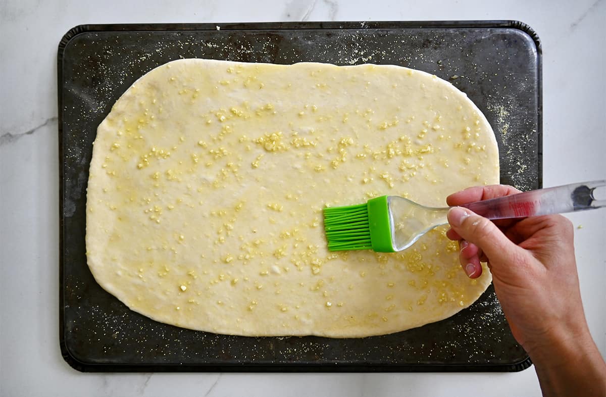 A hand holding a pastry brush spreads a garlic-infused olive oil atop pizza dough on an inverted baking sheet.