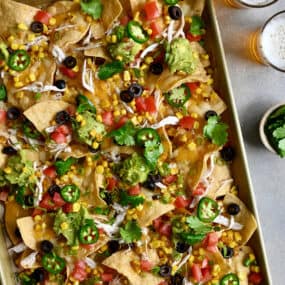 Sheet pan nachos loaded with sliced jalapeños, diced tomatoes, black olives and guacamole next to two glasses filled with light beer.