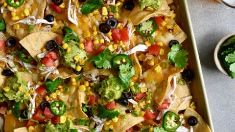 Sheet pan nachos loaded with sliced jalapeños, diced tomatoes, black olives and guacamole next to two glasses filled with light beer.