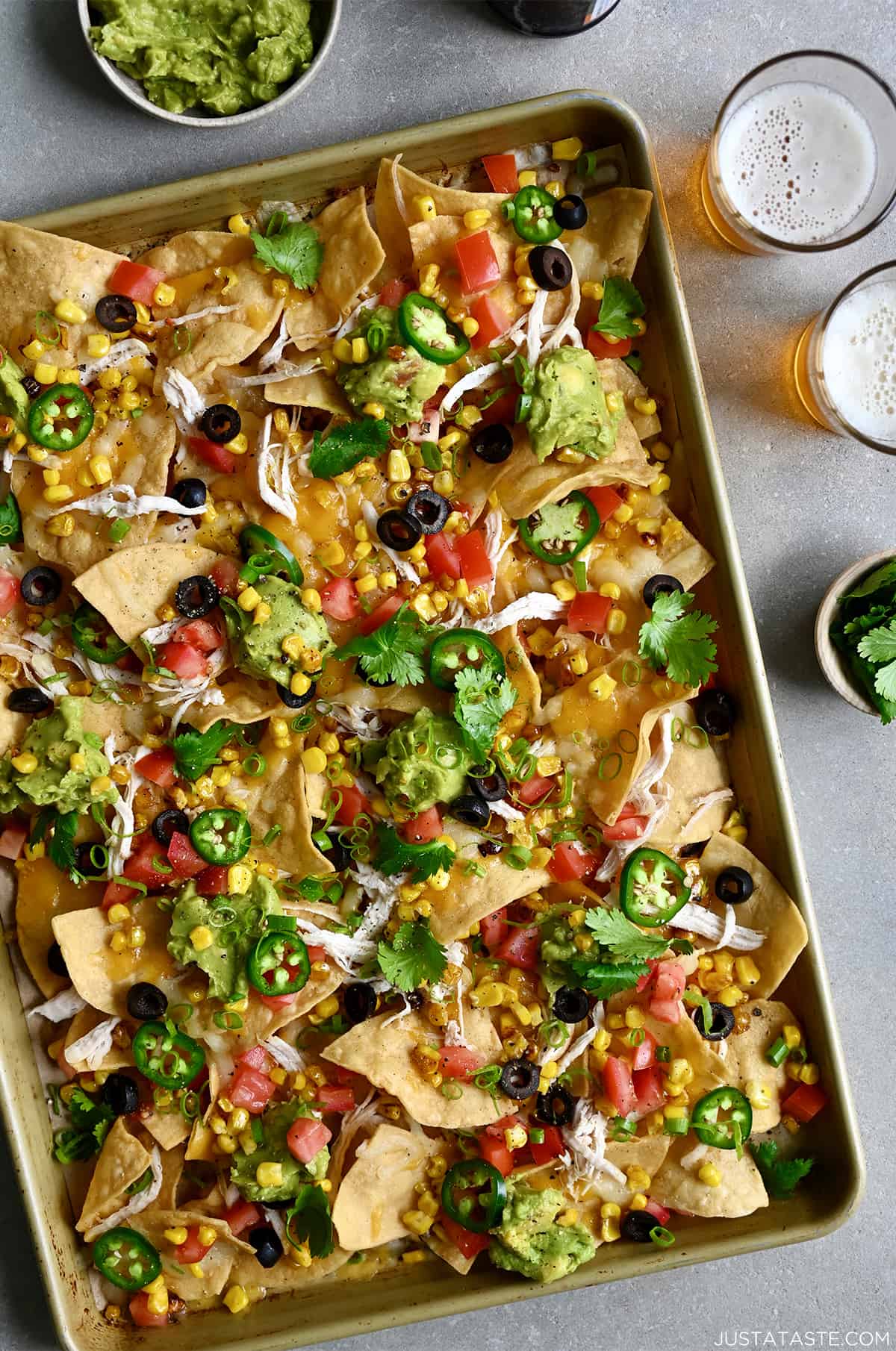 Sheet pan nachos loaded with sliced jalapeños, diced tomatoes, black olives and guacamole next to two glasses filled with light beer.