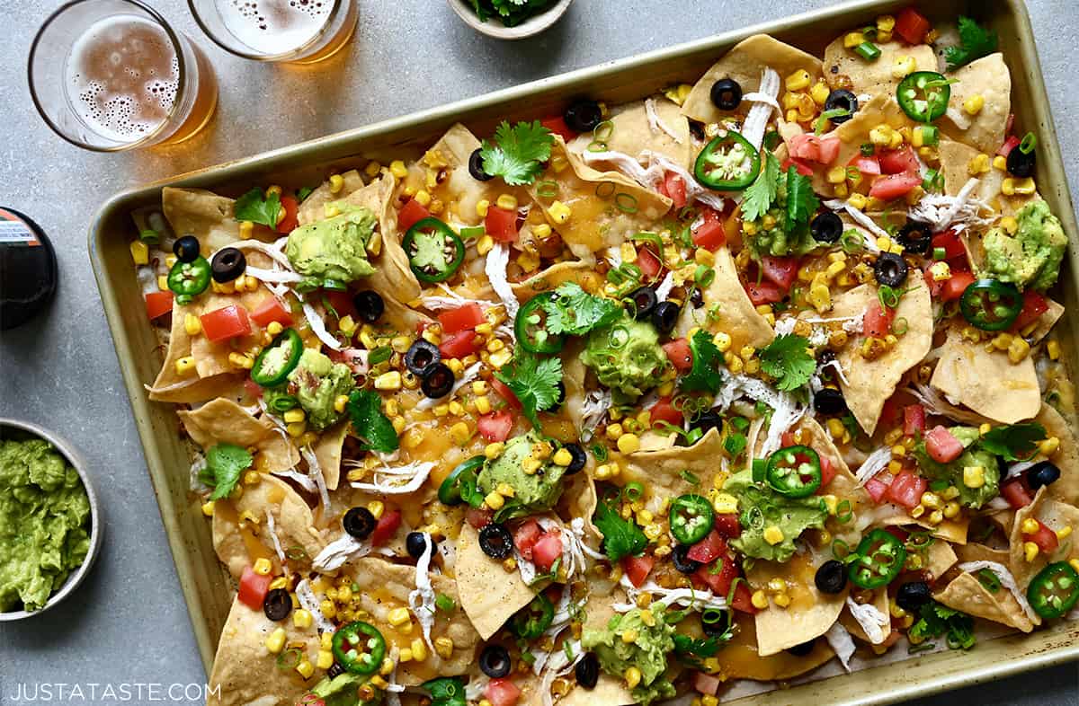 A top-down view of sheet pan nachos loaded with sliced jalapeños, black olives, corn kernels, diced tomatoes, shredded cheese and shredded chicken.