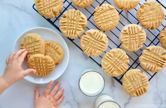 Homemade peanut butter cookies atop a wire cooling rack next to two glasses filled with milk and a child's hands reaching for plate of cookies 