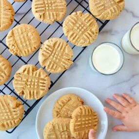 Child's hand reaching for Soft and Chewy Peanut Butter Cookies on white plate next to glasses of milk and wire cooling rack with cookies