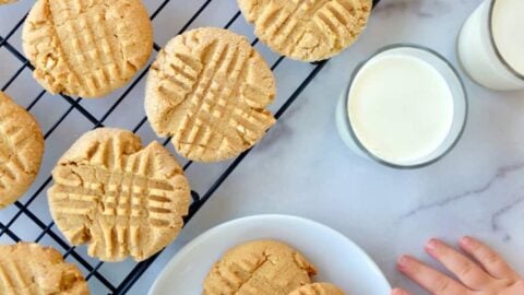 Child's hand reaching for Soft and Chewy Peanut Butter Cookies on white plate next to glasses of milk and wire cooling rack with cookies