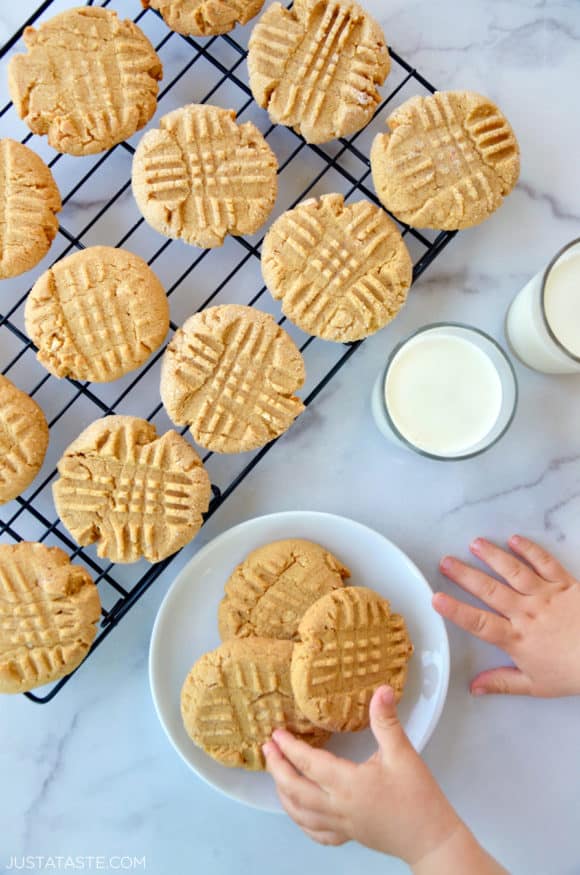 Child's hand reaching for Soft and Chewy Peanut Butter Cookies on white plate next to glasses of milk and wire cooling rack with cookies