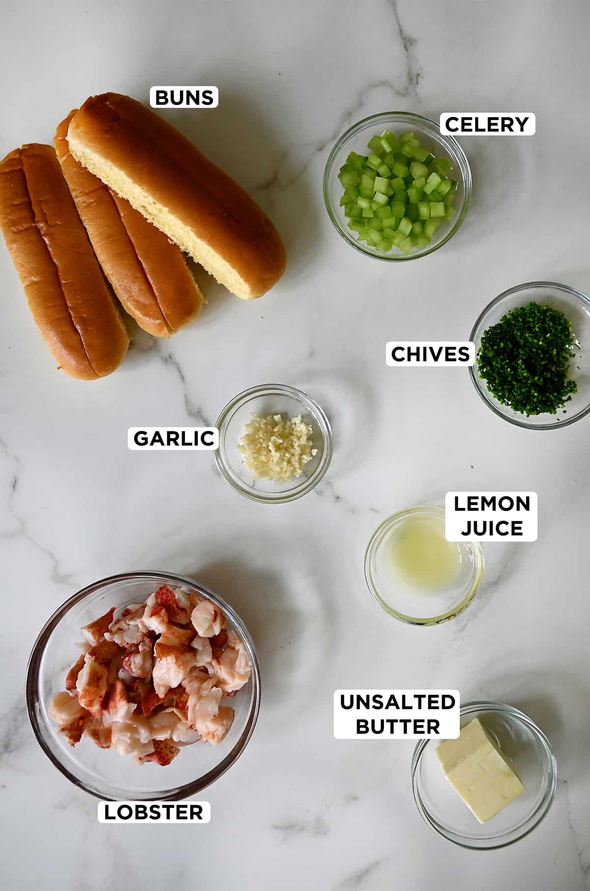 Split-top buns and small clear bowls containing chopped celery, chives, lemon juice, unsalted butter, cooked lobster and minced garlic.