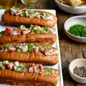 Four lobster rolls on a white serving plate next to a small bowl containing black pepper and a small bowl containing chopped fresh chives.
