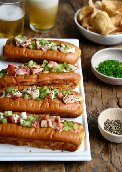 Four lobster rolls on a white serving plate next to a small bowl containing black pepper and a small bowl containing chopped fresh chives.