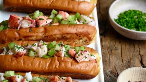 Four lobster rolls on a white serving plate next to a small bowl containing black pepper and a small bowl containing chopped fresh chives.