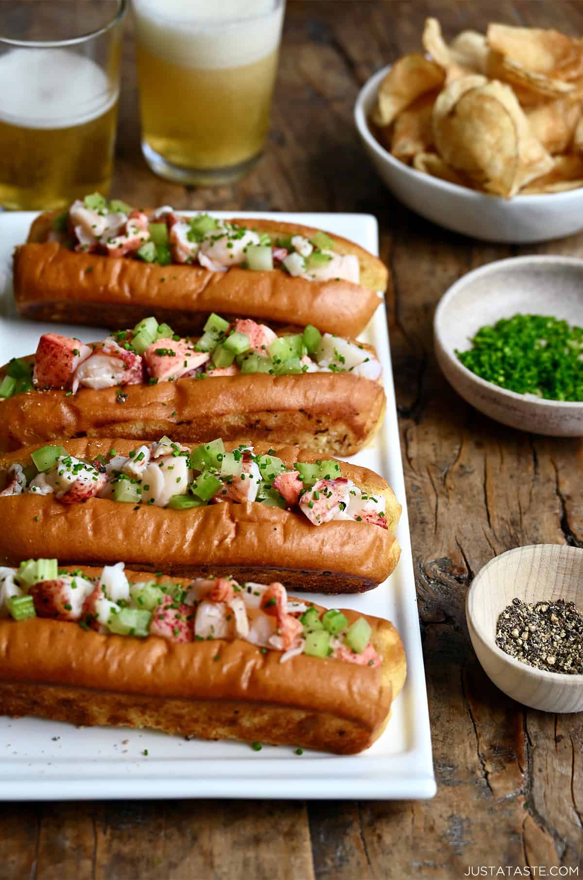 Four lobster rolls on a white serving plate next to a small bowl containing black pepper and a small bowl containing chopped fresh chives.