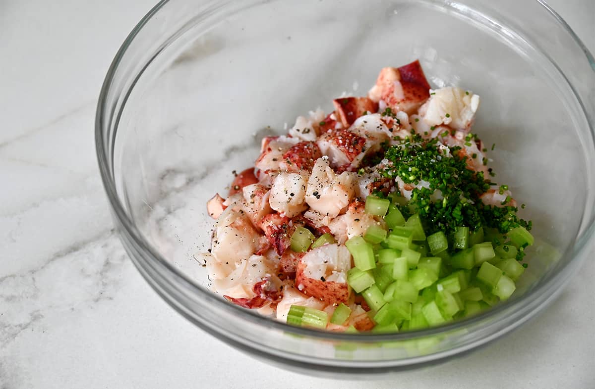 A clear bowl containing cooked lobster meat, chopped celery, chopped fresh chives, salt and pepper.