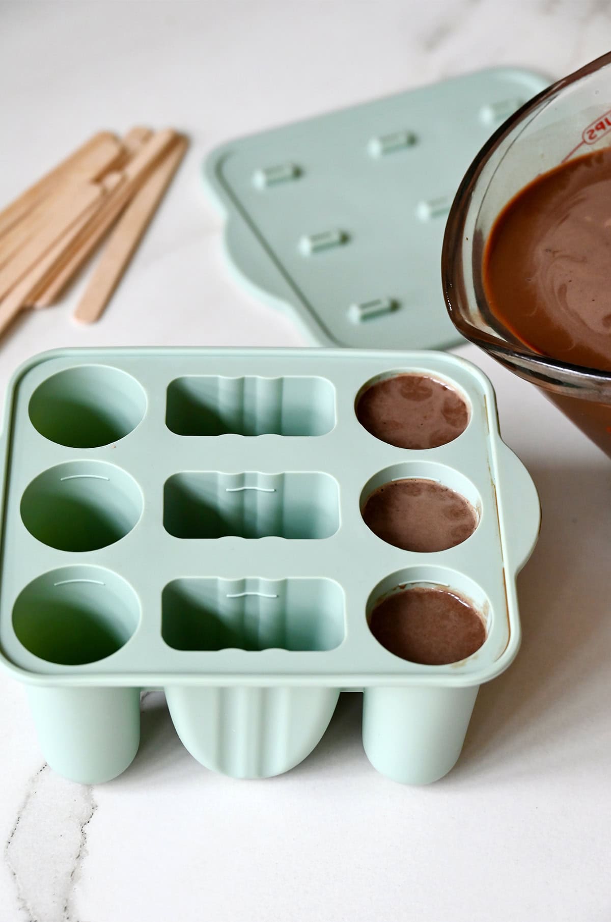 Pouring a homemade fudgesicles mixture from a liquid measuring cup into silicone popsicle molds.