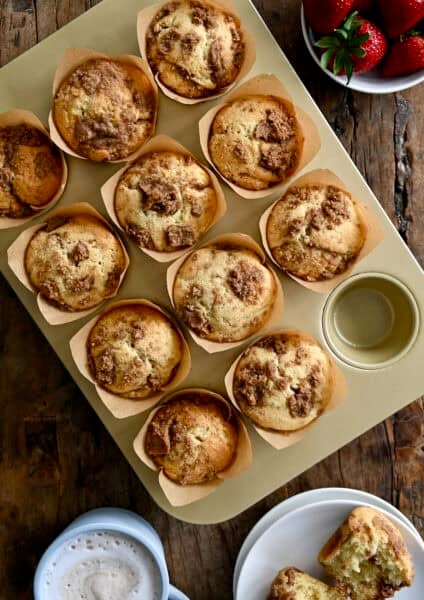 Coffee cake muffins with cinnamon streusel in a muffin pan, with one on a plate, sliced open and slathered with butter.