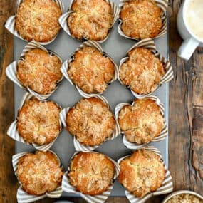 A top-down view of Sour Cream Coffee Cake Muffins with Streusel in a muffin tin next to a latte