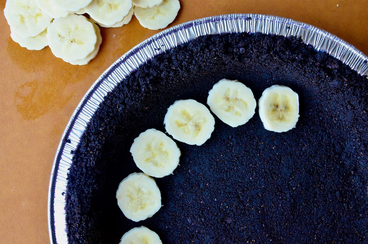 A handful of banana slices on a chocolate cookie pie crust. More banana slices sit beside the pie plate.
